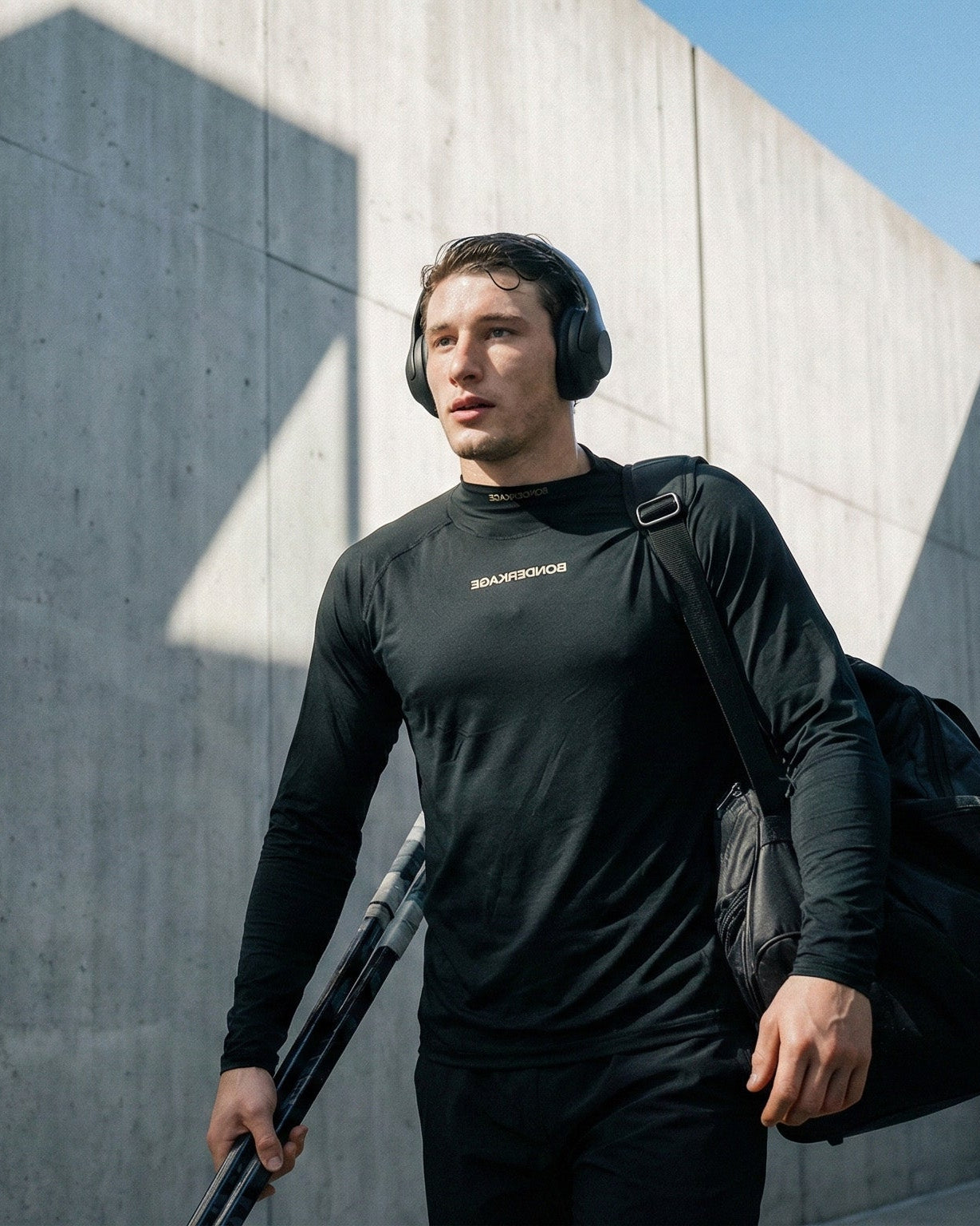 Man in black wearing Bonderkage wear with a backpack and headphones, standing against a concrete wall